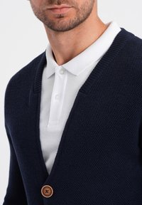 Cárdigan de punto azul marino con diseño de pico, que presenta un cierre de botón de madera y una camisa blanca con cuello debajo.