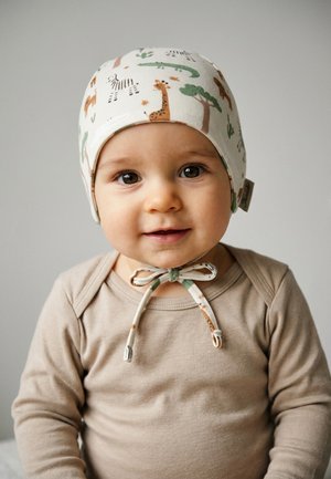 Smiling baby wearing a beige long-sleeve top and a white hat with animal and tree prints, tied under the chin with a bow.