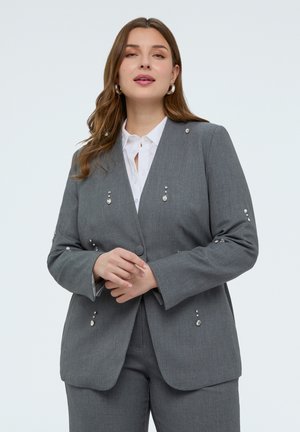 Vrouw draagt een grijs op maat gemaakt pak met wit overhemd, versierd met kleine glanzende versieringen, staand met handen gevouwen voor zich.