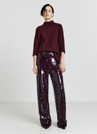 Maglione a collo alto in maglia bordeaux con maniche a coste, abbinato a pantaloni a vita alta in paillettes di colore viola scuro, caratterizzati da una texture lucida.
