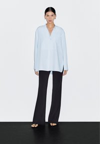 Lichtblauw overhemd met lange mouwen, kraag en knoopsluiting aan de voorkant, gecombineerd met zwarte wijde pijpen broek. Gladde stof en losse pasvorm.