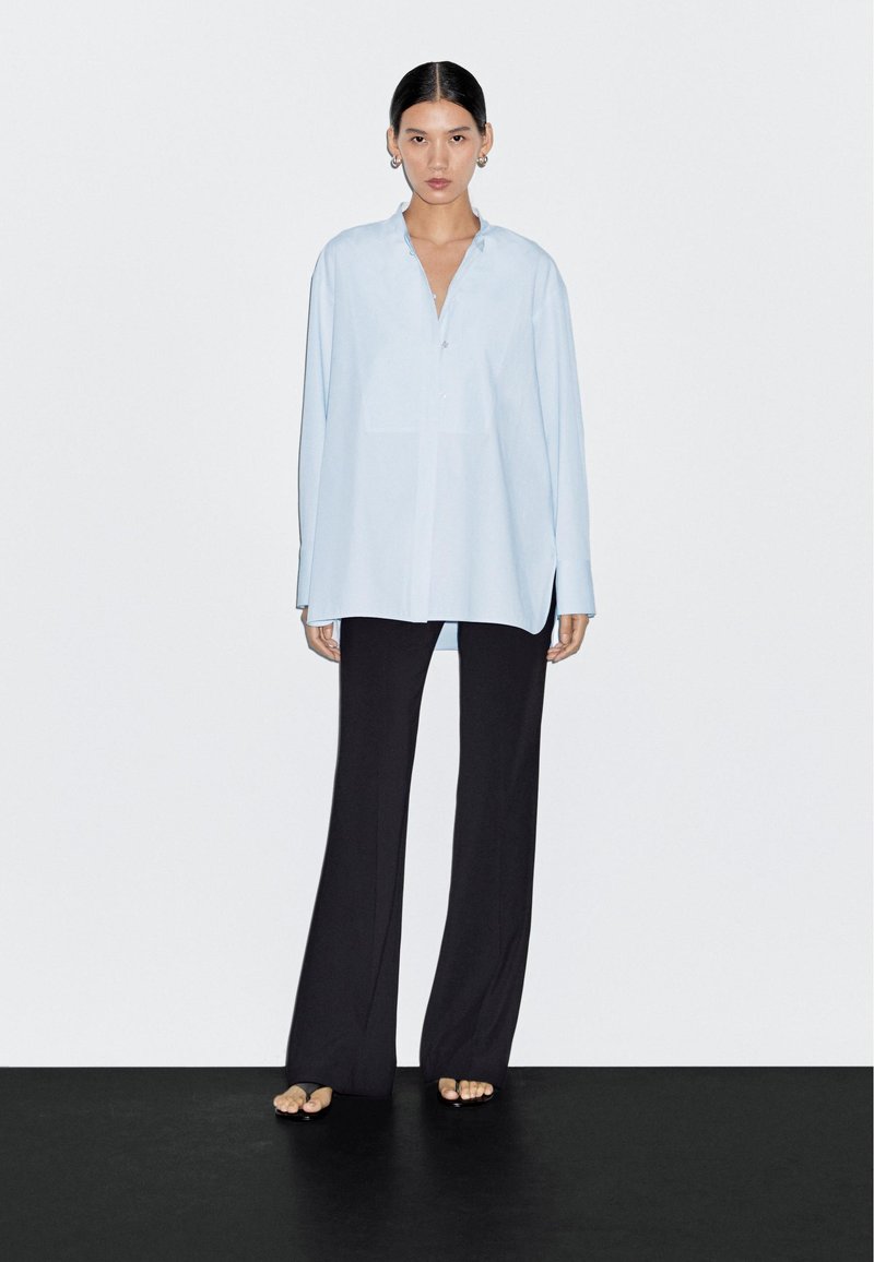 Lichtblauw overhemd met lange mouwen, kraag en knoopsluiting aan de voorkant, gecombineerd met zwarte wijde pijpen broek. Gladde stof en losse pasvorm.