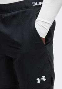Άτομο που φοράει μαύρα σορτσάκια Under Armour με μικρές οπές και λευκό πουκάμισο μακρυμάνικο, το χέρι στην τσέπη.