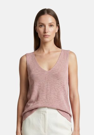 Donna che indossa un top rosa lavorato a maglia senza maniche con scollatura a V e pantaloni bianchi a vita alta, in piedi davanti a uno sfondo chiaro e uniforme.