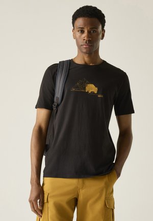Young man with black backpack wearing black t-shirt with yellow truck and mountain design, and mustard cargo pants, standing neutral against gray background.