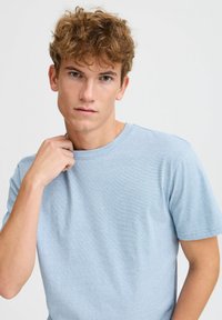 Light blue short-sleeve t-shirt with a round neckline, soft texture, and subtle stripes. Worn by a model with light curly hair.