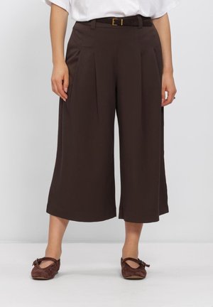 Femme portant un pantalon marron large court avec des plis à l'avant, une ceinture noire, un haut blanc et des chaussures plates marron avec des brides à la cheville.