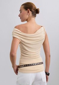 Top beige con los hombros al descubierto y textura recogida, que presenta una silueta ajustada y un cinturón marrón con tornillos metálicos circulares, combinado con pantalones blancos.