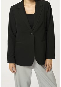 Blazer nero con rever a punta, chiusura a un bottone, due tasche frontali e maniche lunghe, abbinato a pantaloni grigi.