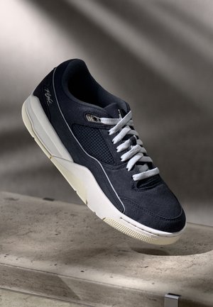 Czarno-biały sportowy sneaker z białymi sznurowadłami, panelami z siateczki i grubą białą podeszwą spoczywający na teksturowanej kamiennej powierzchni.