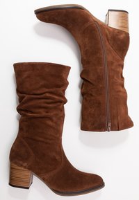 Botas de ante marrón hasta la mitad de la pantorrilla con tacones de bloque apilados, eje holgado y cremallera lateral sobre un fondo blanco.