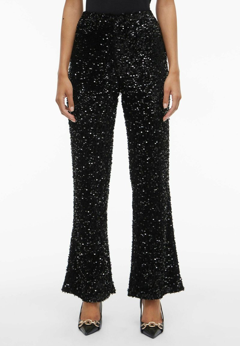 Črne hlače z bleščicami s širokimi nogavicami, teksturirane površine in visokim pasom. Kombinirane s črnimi koničastimi petami z zlato strojno opremo.