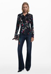 Blusa nera a fiori con maniche lunghe, caratterizzata da motivi floreali multicolori. Abbinata a jeans attillati blu scuro e tacchi a punta neri.