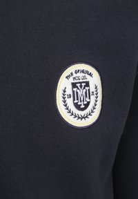 Marine gebreide stof met een geborduurd circulair logo met zwarte en witte elementen, tekst en laurierdetails.