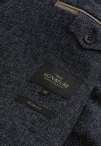 Traje de tejido texturizado en navy, con una etiqueta prominente que dice "next SIGNATURE Premium Tailoring", tamaño 40R y etiqueta de "SLIM FIT".