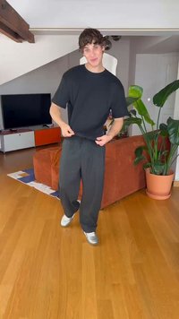 Svart ribbet kortermet skjorte med løstsittende grå bukser, parret med grå sneakers. Rommet har tregulv og en grønn plante.