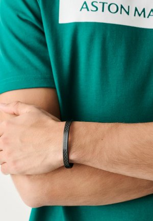 Osoba v zelenom tričku Aston Martin s čiernym textúrovaným náramkom značky Aston Martin na ľavom zápästí, so skríženými rukami.