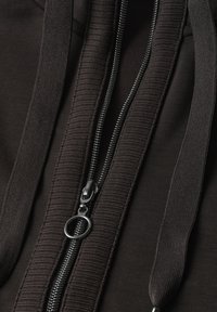 Close-up of a black zipper with a round metal pull on textured fabric and two flat black drawstrings.