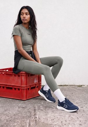 Kvinde iført grønt træningstøj og navy sneakers sidder på stablede røde plastik kasser mod en hvid væg.