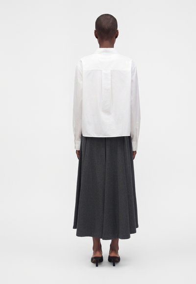 Personne portant une chemise blanche à manches longues, une jupe midi gris foncé et des chaussures noires à talons, debout de dos à la caméra.