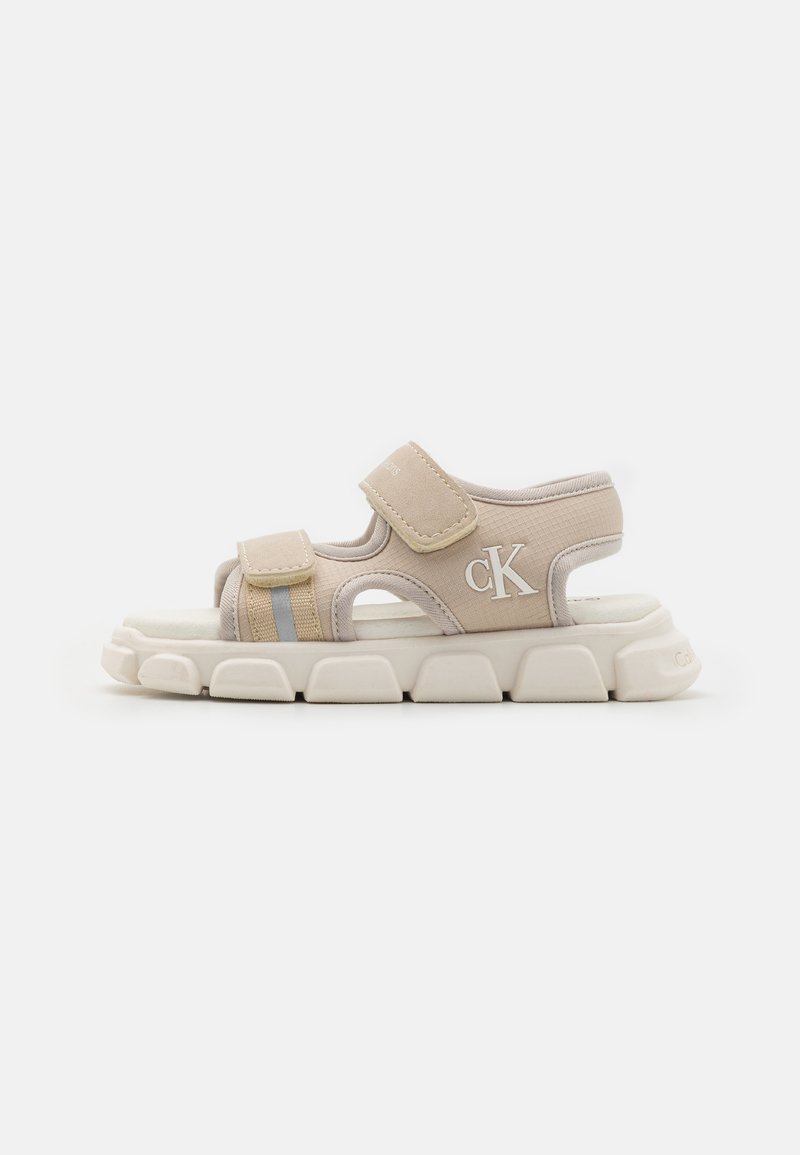 Beige sandaler med borrelåsremmer, teksturert overdel, polstret såle og en uttalt hvit mellomsåle. Har en subtil logo og grå detaljer.