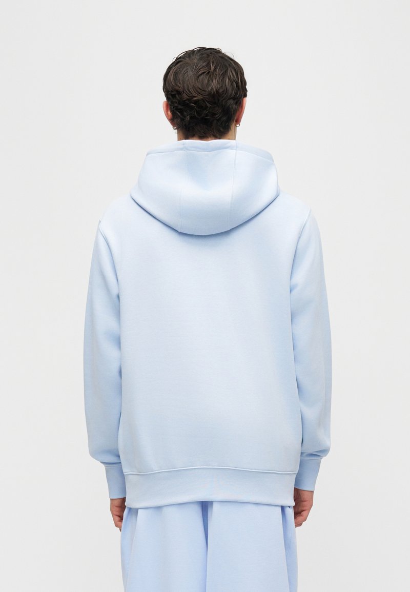 Persona con cabello corto y oscuro, vistiendo una sudadera con capucha azul claro y pantalones a juego, de espaldas contra un fondo liso y claro.