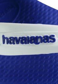 Chanclas Havaianas azules con tira blanca, con texto azul en relieve y una suela texturizada azul con patrones ovalados. Material de goma durable.