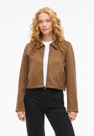 Hosszú, göndör szőke hajú nő barna, cipzáros dzsekit visel fehér ing felett, fekete nadrággal, fehér háttér előtt állva.