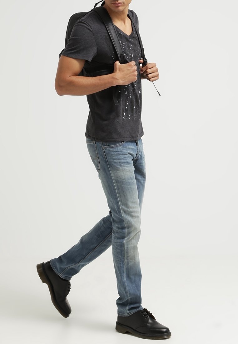 Camiseta negra de cuello en V con un sutil patrón de salpicaduras, jeans de mezclilla azul claro y zapatos negros con cordones; modelo llevando una mochila.