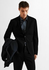 Mlad moški v črnem krilu in vzorčeni temno modri srajci drži črno jakno in gleda navzdol pred enobarvno belo ozadje.