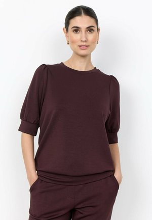 Vrouw met donker haar in een kastanjebruine top met pofmouwen en bijpassende broek, staand met handen in de zakken tegen een effen witte achtergrond.