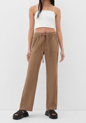 Femme portant un pantalon large marron taille haute avec cordon à la taille, un débardeur blanc court et des sandales plateformes noires.