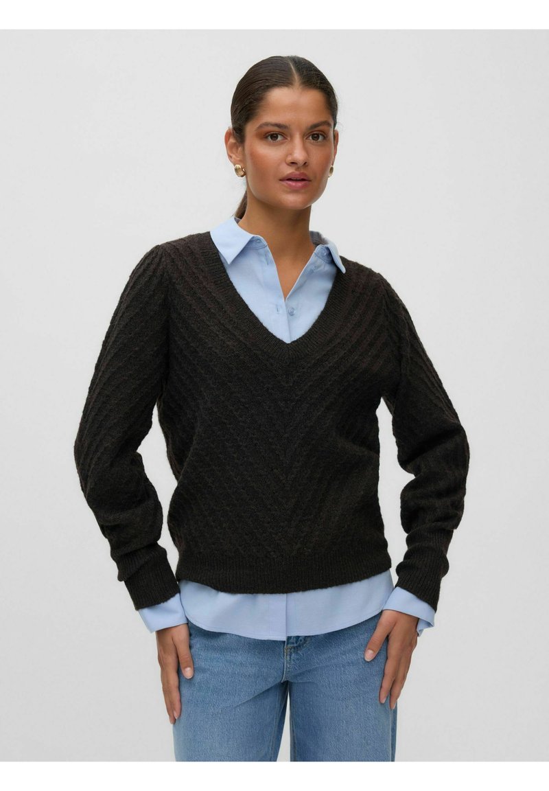 Femme portant un pull en maille marron foncé à col en V sur une chemise bleu clair à col et un jean bleu, debout devant un fond uni.