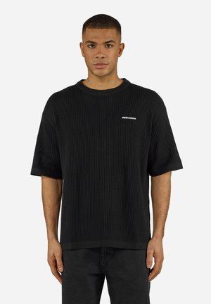 Muž v čiernom žíhanom tričku s krátkym rukávom s logom "PEGADOR" na hrudi, stojaci pred jednoduchým bielym pozadím.