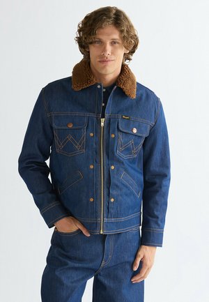 Mann trägt blaue Jeansjacke mit braunem Lammfellkragen und passenden Jeanshosen, steht vor einem schlichten hellen Hintergrund.