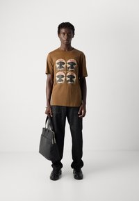 Junger Mann in braunem Grafik-T-Shirt und schwarzen Hosen, der eine schwarze Ledertasche hält, steht vor einem schlichten weißen Hintergrund.