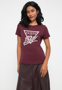 Vrouw met donker haar die een maroon Guess t-shirt draagt met een wit driehoekig logo en een donker leren rok, staande tegen een effen achtergrond.