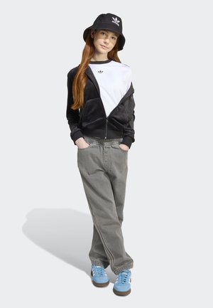 Veste zippée en velours noir sur T-shirt blanc, pantalon large gris et baskets bleues. Chapeau seau noir avec logo, le modèle a de longs cheveux roux.