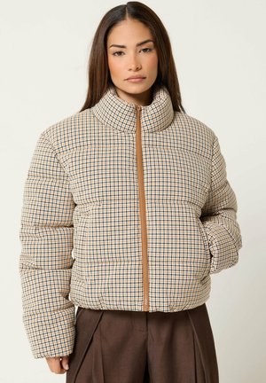 Femme portant une doudoune beige à carreaux avec un col montant et un pantalon marron, debout avec une main dans la poche sur un fond uni.
