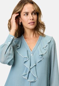 Blouse bleu clair avec un col en V, ornée de détails froncés et d'un galon blanc. Manches longues avec poignets à boutons. Tissu texturé.