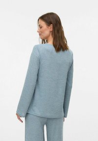 Maglietta azzurra chiara a maniche lunghe con tessuto testurizzato e colletto rotondo, abbinata a pantaloni larghi coordinati. La vista posteriore mostra una semplicità elegante.