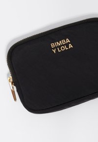 Neceser negro de nylon con cremallera y logo dorado que dice "BIMBA Y LOLA." Presenta una superficie texturizada y un detalle de cremallera en contraste.