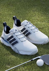 Chaussures de golf blanches avec des rayures noires sur l'herbe à côté d'un club de golf et d'une balle de golf.