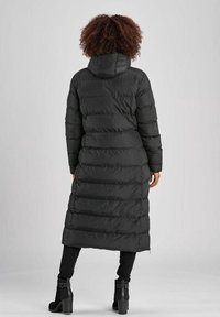 Lange zwarte donsjas met capuchon, voorzien van doorgestikt ontwerp, verborgen ritssluiting en aansluitende manchetten. Draagt over strakke zwarte broek en laarzen.