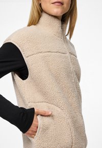 Vestă din fleece bej cu guler înalt, închidere cu fermoar și buzunare laterale. Prezintă o suprafață texturată și o margine contrastantă de-a lungul muchiilor.