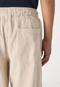 Beżowe spodnie sztruksowe o prążkowanej fakturze, z elastycznym pasem i naszywaną kieszenią na plecach, z widoczną metką marki z boku.