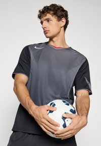 Young man in black-gray sportswear holding a white and blue soccer ball, looking sideways against a plain light background.