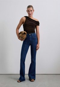 Top marrone a spalla unica, abbinato a jeans a vita alta blu scuro a zampa d'elefante. Modella tiene una borsa a pochette color sabbia. Tessuto morbido e design aderente.