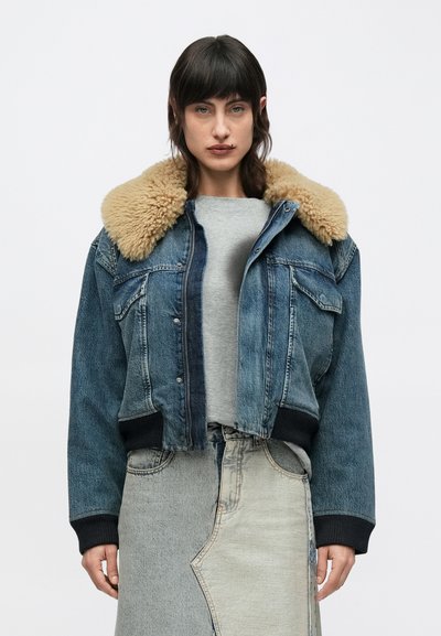 Νεαρή γυναίκα φορώντας ένα μπλε τζιν μπουφάν με μπεζ γιακά από shearling και μια δίχρωμη γκρι τζιν φούστα, στέκεται μπροστά σε λευκό φόντο.