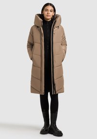 Kvinde iført en lang beige pufferfrakke med høj krave, sort ribbet outfit og sorte klodsede støvler, stående mod en lys baggrund.